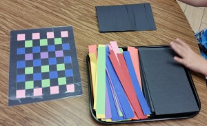 A sample of a finished placemat, pre-cut strips for weaving, and folded black paper with lines